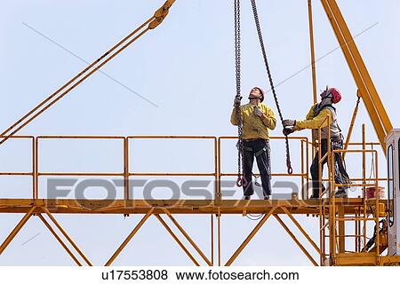 Close up of horizontal part of yellow crane near cabin with two men in yellow jackets, and red safety helmets, working with hanging chains and hooks, Umbria, Italy View Large Photo Image Stock Photo - Close up of horizontal part of yellow crane near cabin with two men in yellow jackets, and red safety helmets, working with hanging chains and hooks, Umbria, Italy. Fotosearch
