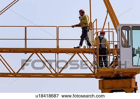 Stock Photo - Close up of horizontal part of yellow crane near cabin with two men in yellow jackets, and red safety helmets, one man walking, the other standing, Umbria, Italy. Fotosearch