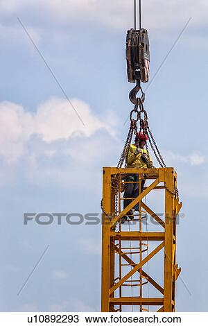 Close up of vertical part of yellow crane with man attached by chains, wearing yellow jackets, and red safety helmets, attaching chains to huge hook above, as he works on crane assembly, Umbria, Italy View Large Photo Image Stock Image - Close up of vertical part of yellow crane with man attached by chains, wearing yellow jackets, and red safety helmets, attaching chains to huge hook above, as he works on crane assembly, Umbria, Italy. Fotosearch