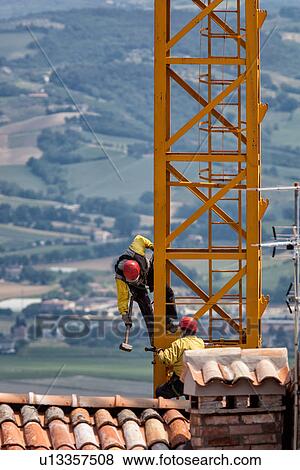 Close up of vertical part of yellow crane with two men attached by chains, wearing yellow jackets, and red safety helmets, hammering as they work on crane assembly, red tiled roof tops visible in foreground, and green valley beyond, Umbria, Italy View Large Photo Image Stock Photo - Close up of vertical part of yellow crane with two men attached by chains, wearing yellow jackets, and red safety helmets, hammering as they work on crane assembly, red tiled roof tops visible in foreground, and green valley beyond, Umbria, Italy. Fotosearch