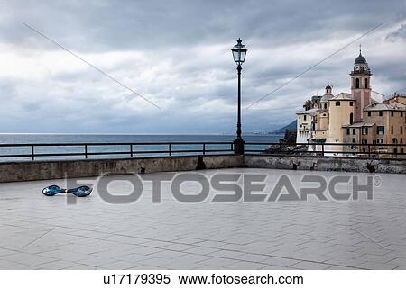 Stock Photography - Deserted terrace of Camogli at dusk with single abandoned skateboard, church tower and buildings, coastline beyond the iron railing, Camogli, Liguria, Italy. Fotosearch