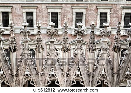 Stock Image - Detail of Milan Duomo rooftop: series of rooftop pinnacles, elaborate stone carving, with palazzo facade beyond, desaturated view, Milan Italy. Fotosearch