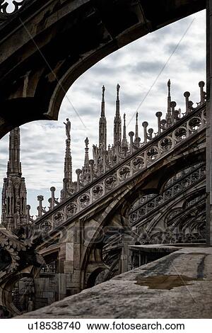 Stock Image - Detail of Milan Duomo rooftop: series of rooftop pinnacles and flying buttresses, elaborate stone carving, against sky, desaturated view, Milan Italy. Fotosearch