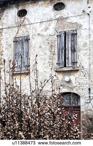 Stock Image - Detail of old stucco house with crumbling facade, ancient wooden shutters over window, typical style, with dying bush in foreground, Emilia Romagna, Italy. Fotosearch