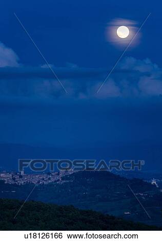 Stock Photograph - Full moon rising over hilltop town of Todi in blue twilight with gentle clouds over Tiber valley, hills beyond, Umbria, Italy. Fotosearch