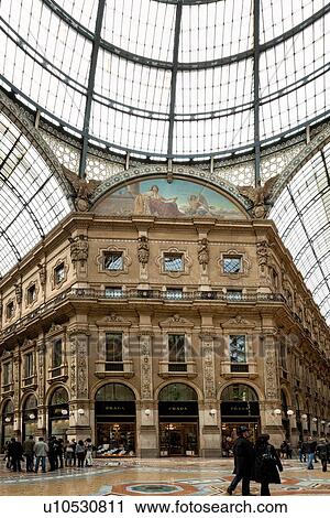 Stock Image - Gallerica Vittorio Emanuele: detail view of central mall area, people shopping, glass and cast iron central vaulted dome and arched roof with frescoed and stone carving decoration, Milan Italy. Fotosearch