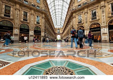 Stock Image - Gallerica Vittorio Emanuele: detail view of central mall area, people shopping, glass and cast iron arched roof with stone carving decoration, Milan Italy. Fotosearch