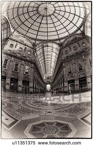 Stock Photography - Gallerica Vittorio Emanuele: detail view of central mall area, people shopping, glass and cast iron arched roof with stone carving decoration, mosaic flooring, monochrome view, Milan Italy. Fotosearch