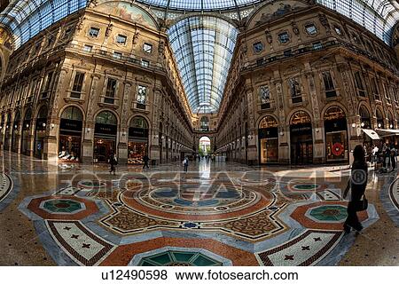 Gallerica Vittorio Emanuele: wide angle view of central mall area, people shopping, glass and cast iron arched roof with stone carving decoration, mosaic flooring, Milan Italy View Large Photo Image Stock Photo - Gallerica Vittorio Emanuele: wide angle view of central mall area, people shopping, glass and cast iron arched roof with stone carving decoration, mosaic flooring, Milan Italy. Fotosearch