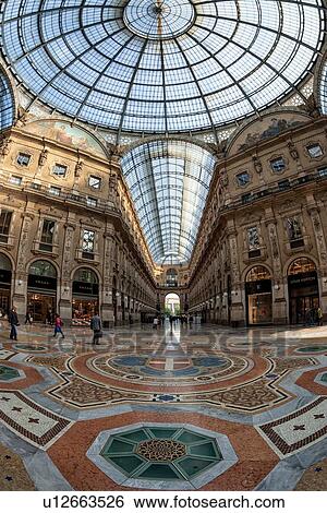 Stock Photograph - Gallerica Vittorio Emanuele: wide angle view of central mall area, people shopping, glass and cast iron arched roof with stone carving decoration, central glass dome, mosaic flooring, Milan Italy. Fotosearch