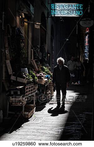 Genoa Centro Storico: narrow alley with fruit and vegetable stand in chiaroscuro light, with man walking, strong shadows, Genova, Italy View Large Photo Image Stock Image - Genoa Centro Storico: narrow alley with fruit and vegetable stand in chiaroscuro light, with man walking, strong shadows, Genova, Italy. Fotosearch