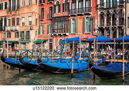 Stock Image - Grand Canal: people in open cafe, ornately decorated palazzi, wooden poles with moored gondolas, late afternoon golden light, Venice, Italy. Fotosearch