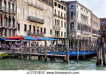 Stock Image - Grand Canal: people in open cafe, ornately decorated palazzi, wooden poles with moored gondolas, late afternoon golden light, Venice, Italy. Fotosearch