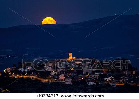 Stock Photo - Huge golden full moon peeping over hills beyond medieval hilltop town of MonteCastello di Vibio with night lights at twilight, and blue sky and hills beyond, Umbria, Italy. Fotosearch