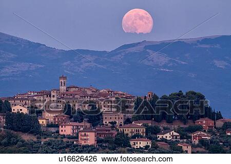 Stock Photograph - Huge pink full moon peeping over hills and cloud beyond medieval hilltop town of MonteCastello di Vibio at twilight, with blue sky, Umbria, Italy. Fotosearch
