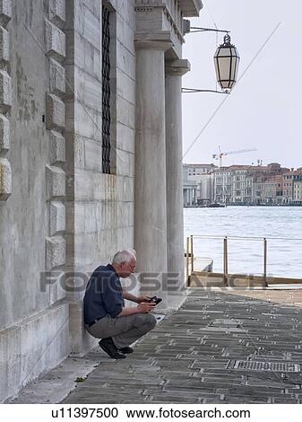 Stock Image - Man reading, crouched alongside building of San Giorgio Maggiore with basin of San Marco beyond, Venice, Italy. Fotosearch