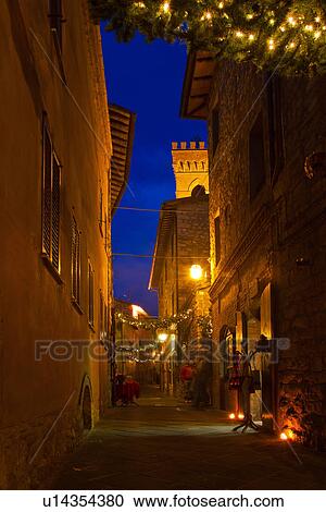 Stock Image - Medieval hilltop town of MonteCastello di Vibio: street at dusk, with night lights, and Christmas garlands strung between the old stone houses, and shops, Umbria, Italy. Fotosearch