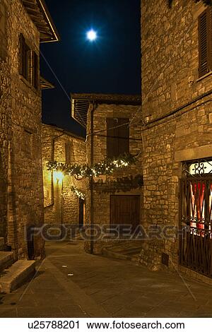 Stock Image - Medieval hilltop town of MonteCastello di Vibio: street at night, lit by night lights and full moon, with Christmas garlands strung between the old stone houses, Umbria, Italy. Fotosearch