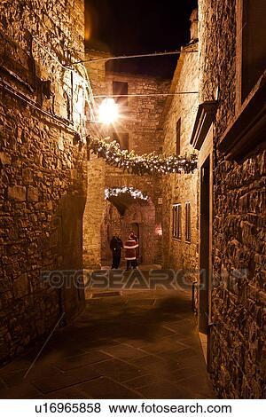 Stock Photo - Medieval hilltop town of MonteCastello di Vibio: street at night, with night lights, and Christmas garlands strung between the old stone houses, "Santa Claus" and two other people, Umbria, Italy. Fotosearch