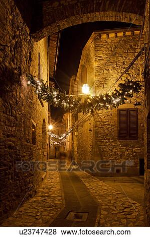 Stock Photo - Medieval hilltop town of MonteCastello di Vibio: street at night, with night lights, and Christmas garlands strung between the old stone houses, motion blur of people, Umbria, Italy. Fotosearch