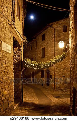 Stock Photograph - Medieval hilltop town of MonteCastello di Vibio: street at night, lit by night lights and full moon, with Christmas garlands strung between the old stone houses, Umbria, Italy. Fotosearch