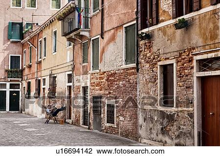 Stock Image - Morning Papers: single man with feet up against the wall, sitting reading the paper in quiet alley with red brick, and painted stucco facade, Venice, Italy. Fotosearch