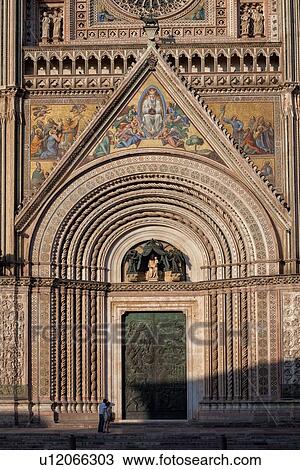 Orvieto Duomo - detail of facade with intricate stone carving, twisted columns with inlaid mosaic panels around bronze carved doors steps lwith two tourists standing below, Romanesque-Gothic style, Umbria, Italy View Large Photo Image Stock Image - Orvieto Duomo - detail of facade with intricate stone carving, twisted columns with inlaid mosaic panels around bronze carved doors steps lwith two tourists standing below, Romanesque-Gothic style, Umbria, Italy. Fotosearch