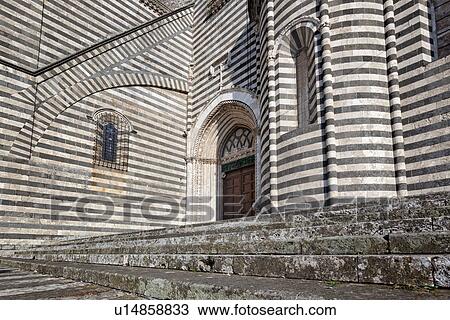 Stock Image - Orvieto Duomo - detail of side wall with black and white striped stone, curved walls and decorations causing dramatic patterned effect, steps leading to ornate doorway Romanesque-Gothic style, Umbria, Italy. Fotosearch