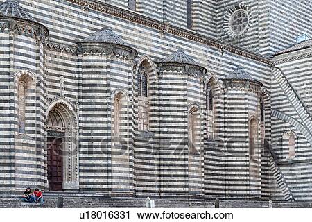Stock Image - Orvieto Duomo - detail of side wall with black and white striped stone, curved walls and decorations causing dramatic patterned effect, steps lwith two people sitting, leading to ornate doorway Romanesque-Gothic style, Umbria, Italy. Fotosearch