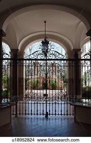 Picture - Palazzo courtyard with arched ceiling, wrought iron hanging lamp, gate leading to open area with wrought iron fencing, and trees beyond, Rome, Italy. Fotosearch