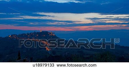 Stock Image - Panorama view of Tiber valley at sunrise with hills silhouetted purple by valley mist, with night lights of hilltop town of MonteCastello di Vibio, against pink and blue sky, Umbria, Italy. Fotosearch
