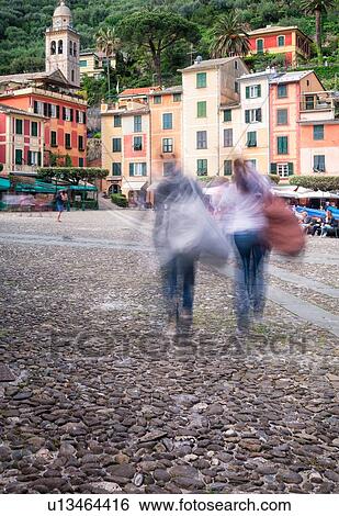 Stock Photograph - Portofino harbour front with motion blur of tourists, brightly coloured apartment buildings, outdoor cafes, church and belltower beyond in lush greenery, Liguria, Italy. Fotosearch