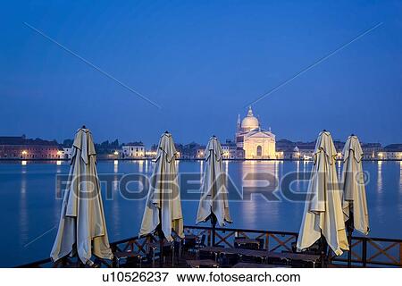 Stock Photo - Redentore church as seen at twilight with lights of the Giudecca, and blue calm water at dawn, closed cafe umbrellas in foregroundVenice, Italy. Fotosearch