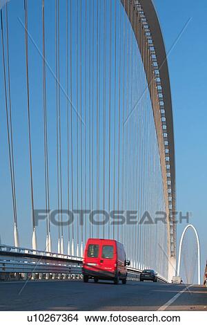 Picture - Reggio Emilia bridge by Calatrava, detail showing curved hoop design and suspension wires with cars passing in foreground, and second bridge beyond, against blue sky in late afternoon, Emilia Romagna, Italy. Fotosearch