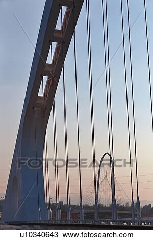 Stock Image - Reggio Emilia bridge by Calatrava, detail showing curved hoop design and suspension wires in foreground, and second bridge beyond, against blue sky at sunset, Emilia Romagna, Italy. Fotosearch