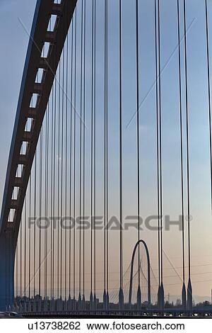 Stock Image - Reggio Emilia bridge by Calatrava, detail showing curved hoop design and suspension wires in foreground, and second bridge beyond, against blue sky at sunset, Emilia Romagna, Italy. Fotosearch