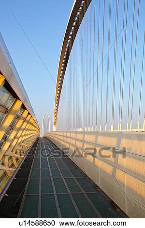 Stock Image - Reggio Emilia bridge by Calatrava, detail showing curved hoop design and suspension wires, with pedestrian sidewalk, second bridge beyond, against blue sky at sunset, Emilia Romagna, Italy. Fotosearch