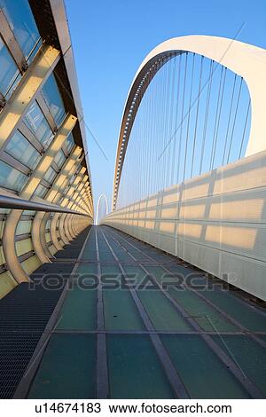 Stock Image - Reggio Emilia bridge by Calatrava, detail showing curved hoop design and suspension wires, with pedestrian sidewalk, second bridge beyond, against blue sky at sunset, Emilia Romagna, Italy. Fotosearch