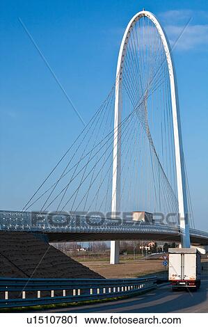 Stock Image - Reggio Emilia bridge by Calatrava, showing curved hoop design and suspension wires against blue sky in late afternoon with trucks on and below bridge, Emilia Romagna, Italy. Fotosearch