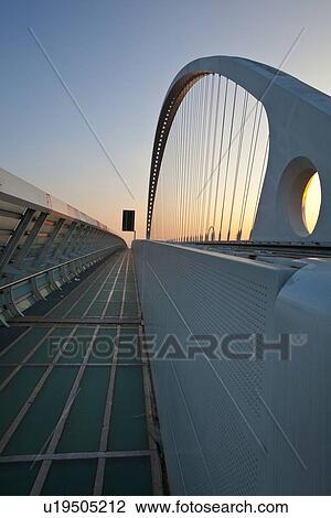 Stock Image - Reggio Emilia bridge by Calatrava, detail showing curved hoop design and suspension wires, with pedestrian sidewalk, second bridge beyond, against blue sky at sunset, Emilia Romagna, Italy. Fotosearch