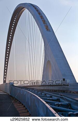 Stock Image - Reggio Emilia bridge by Calatrava, detail showing curved hoop design and suspension wires against blue sky at sunset, Emilia Romagna, Italy. Fotosearch