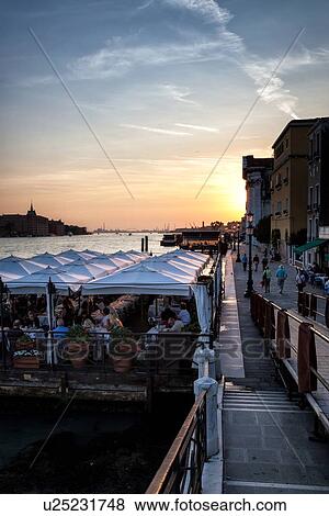 Riva Schiavoni at sunset: steps leading down to open cafe with people dining, view of riva alongside San Marco Basin, with setting sun going behind distant buildings, Venice, Italy View Large Photo Image Stock Photo - Riva Schiavoni at sunset: steps leading down to open cafe with people dining, view of riva alongside San Marco Basin, with setting sun going behind distant buildings, Venice, Italy. Fotosearch