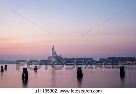 Stock Image - San Giorgio Maggiore as seen at dawn with lights of the Giudecca, and pink calm water reflected from dawn sky, Venice, Italy. Fotosearch
