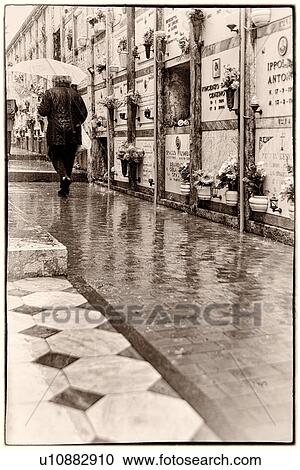 Stock Image - Single woman walking along diagonal path in rain, with umbrella, tombs in background reflected on wet path, Monterosso al Mare, Cinque Terre, Liguria, Italy. Fotosearch