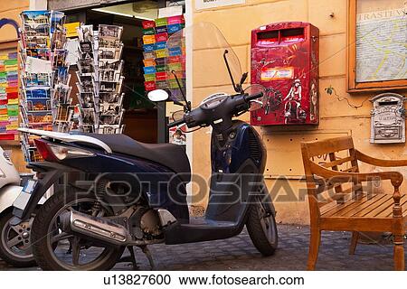 Stock Image - Trastevere street: wooden bench, parked motorbike, and graffiti-covered red letterbox outside shop selling postcards, close up view, Rome, Italy. Fotosearch