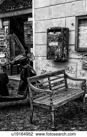 Stock Image - Trastevere street: wooden bench, parked motorbike, and graffiti-covered red letterbox outside shop selling postcards, close up monochrome view, Rome, Italy. Fotosearch