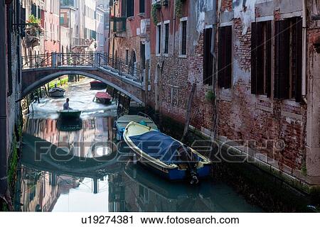 Stock Image - View of canal with motorboat travelling under bridge, other boats moored in early morning sunshine with reflections changing in the wake, Venice, Italy. Fotosearch
