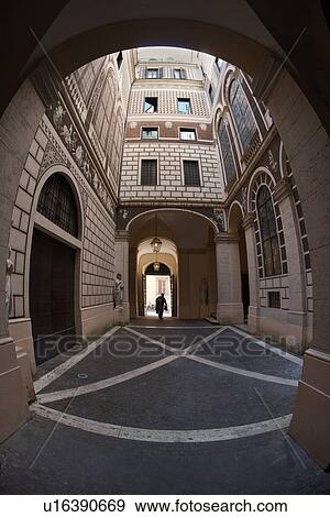 Stock Photo - View of courtyard of palazzo, decorated entirely with faux architectural trompe d'oeil designs of French windows, brick, and balconies, with man entering arched entrance from street, to patterned floor, Rome, Italy. Fotosearch