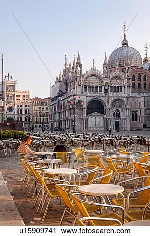 Stock Image - View of the Piazzetta at dawn, with one person sitting in the open cafe, empty yellow chairs, Clock Tower, and Basilica of San Marco, Venice, Italy. Fotosearch