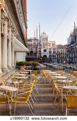Stock Image - View of the Piazzetta at dawn, with one person sitting in the open cafe, empty yellow chairs, Clock Tower, and Basilica of San Marco, Venice, Italy. Fotosearch